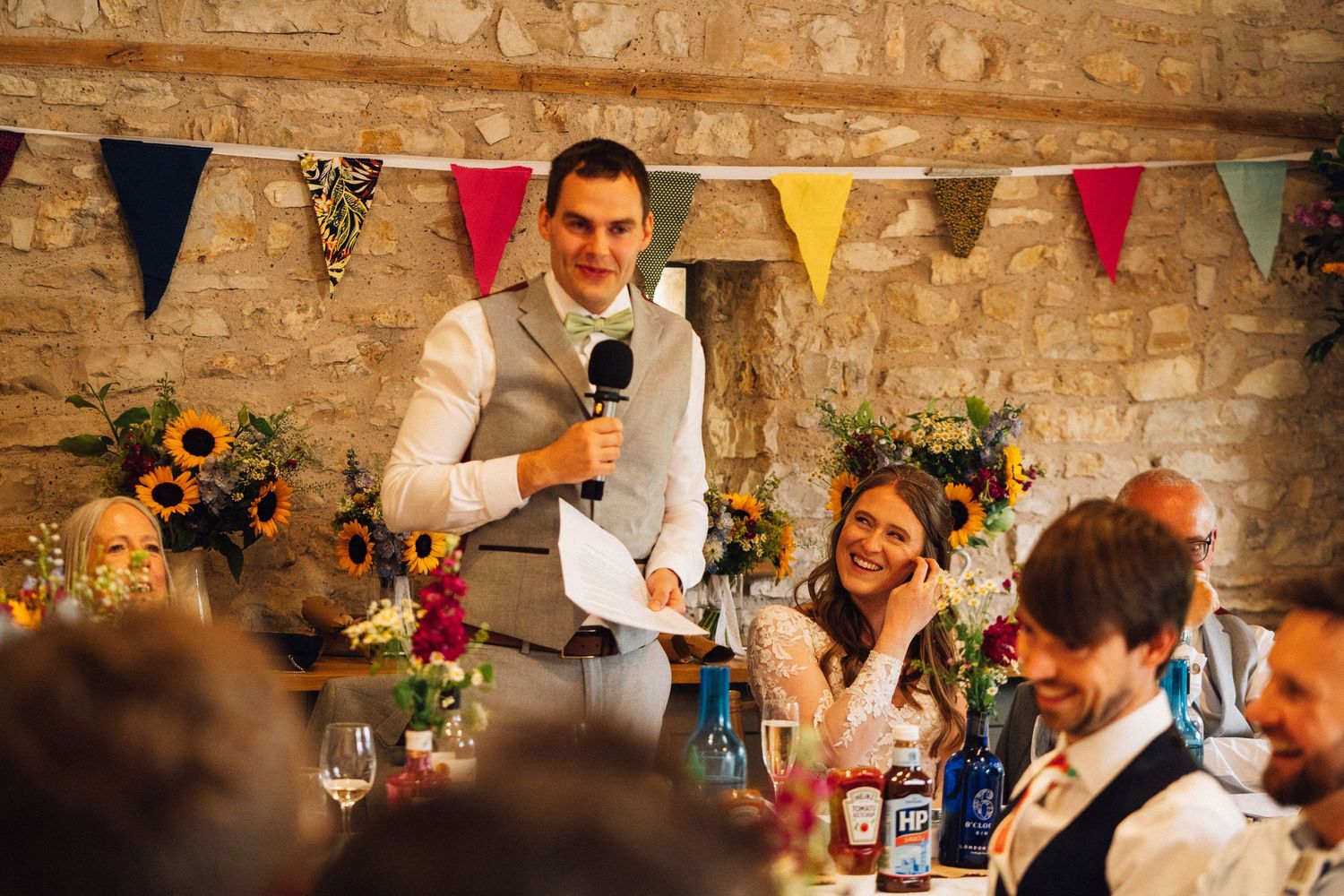 Wedding guests enjoying reception dinner under decorative bunting in stone-walled venue.