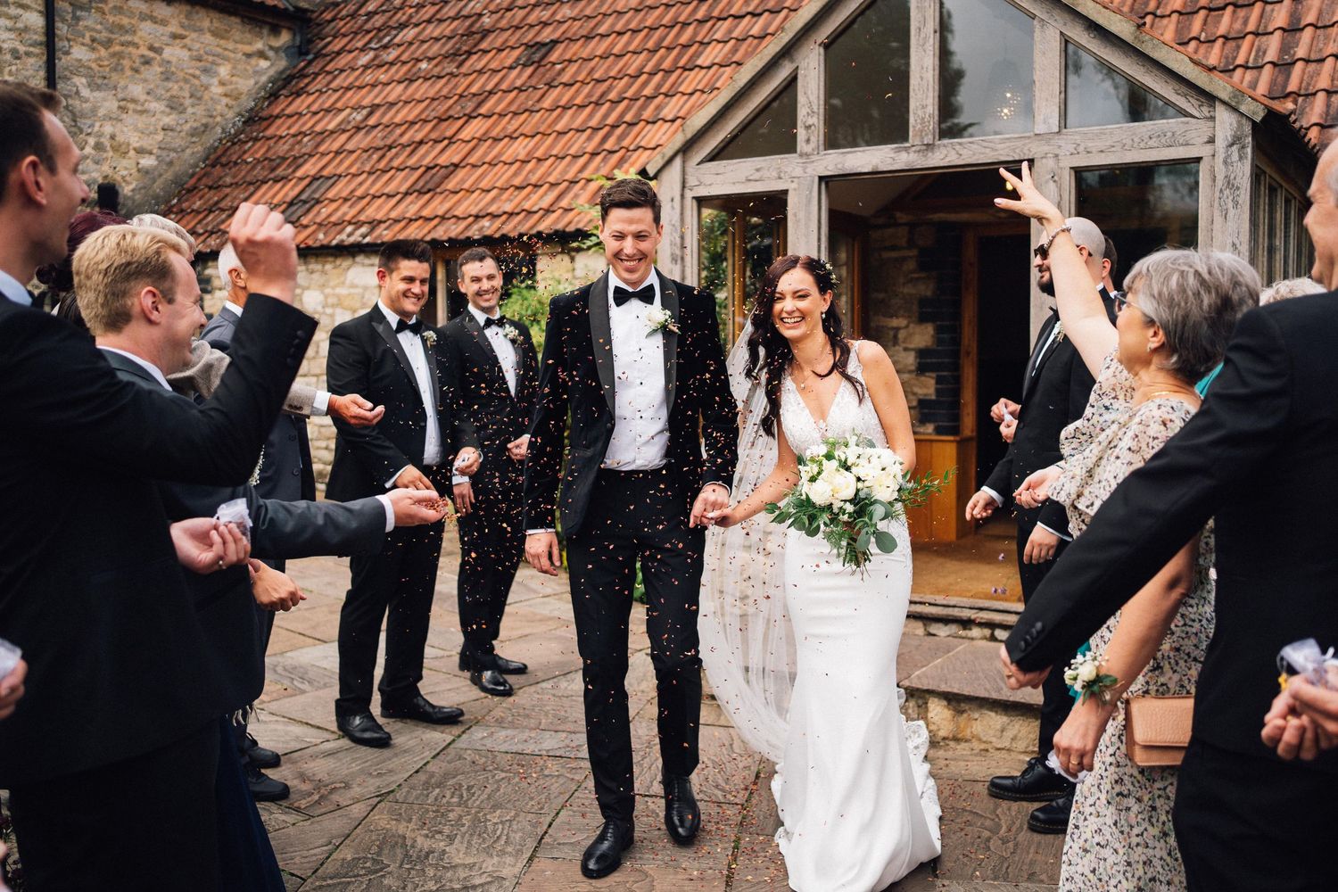 Wedding party exit from rustic stone building with guests throwing confetti.