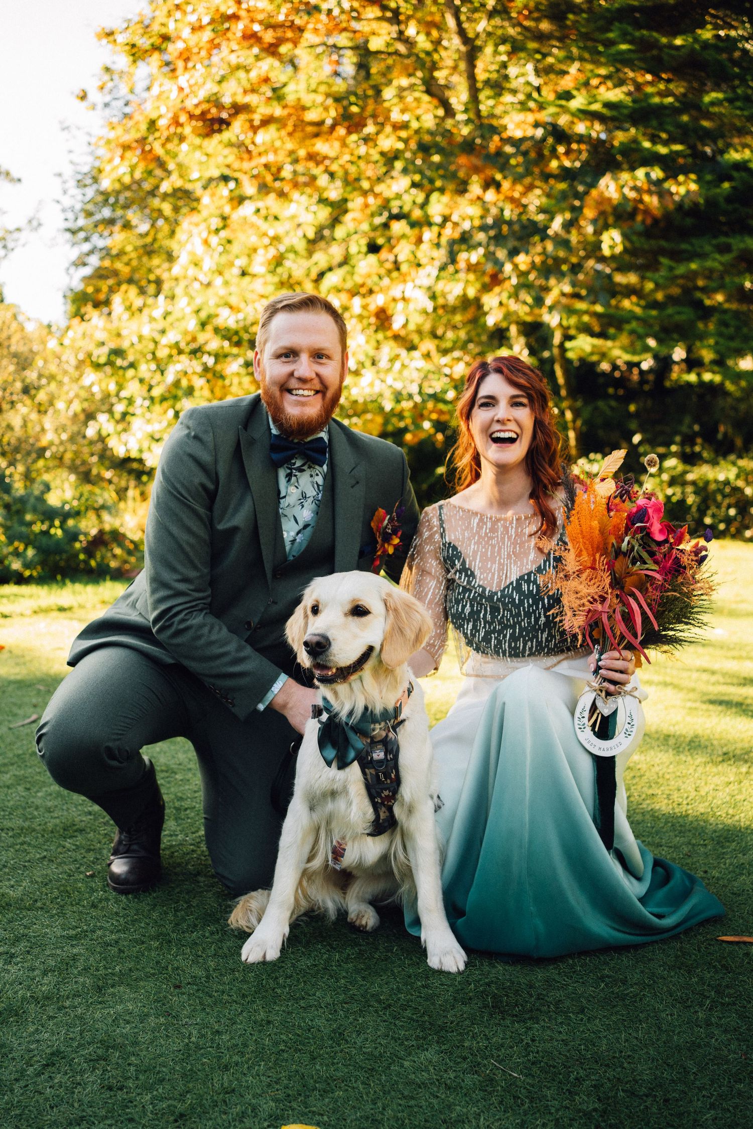 A couple poses with their golden retriever on grass during an autumn wedding photoshoot.