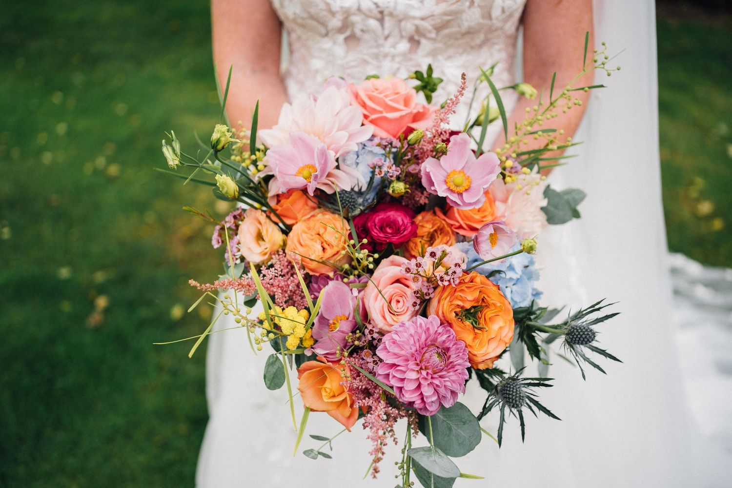 Vibrant bridal bouquet with pink, orange and burgundy flowers mixed with greenery against a grassy background.