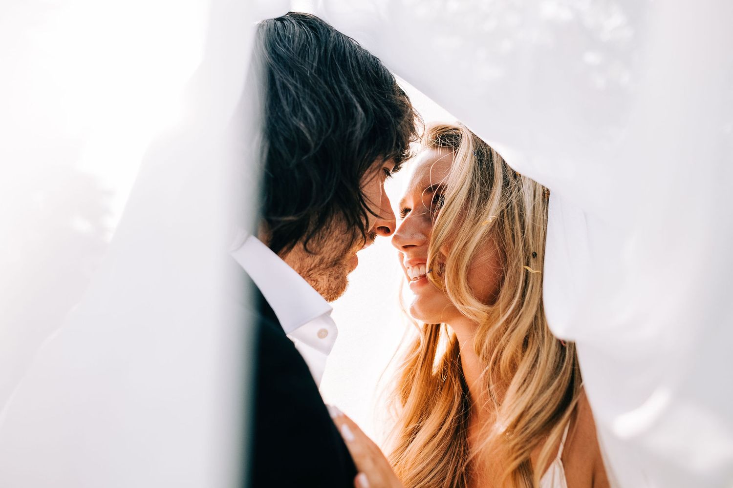 A couple embraces and shares a romantic kiss while standing against a white wall in natural lighting.