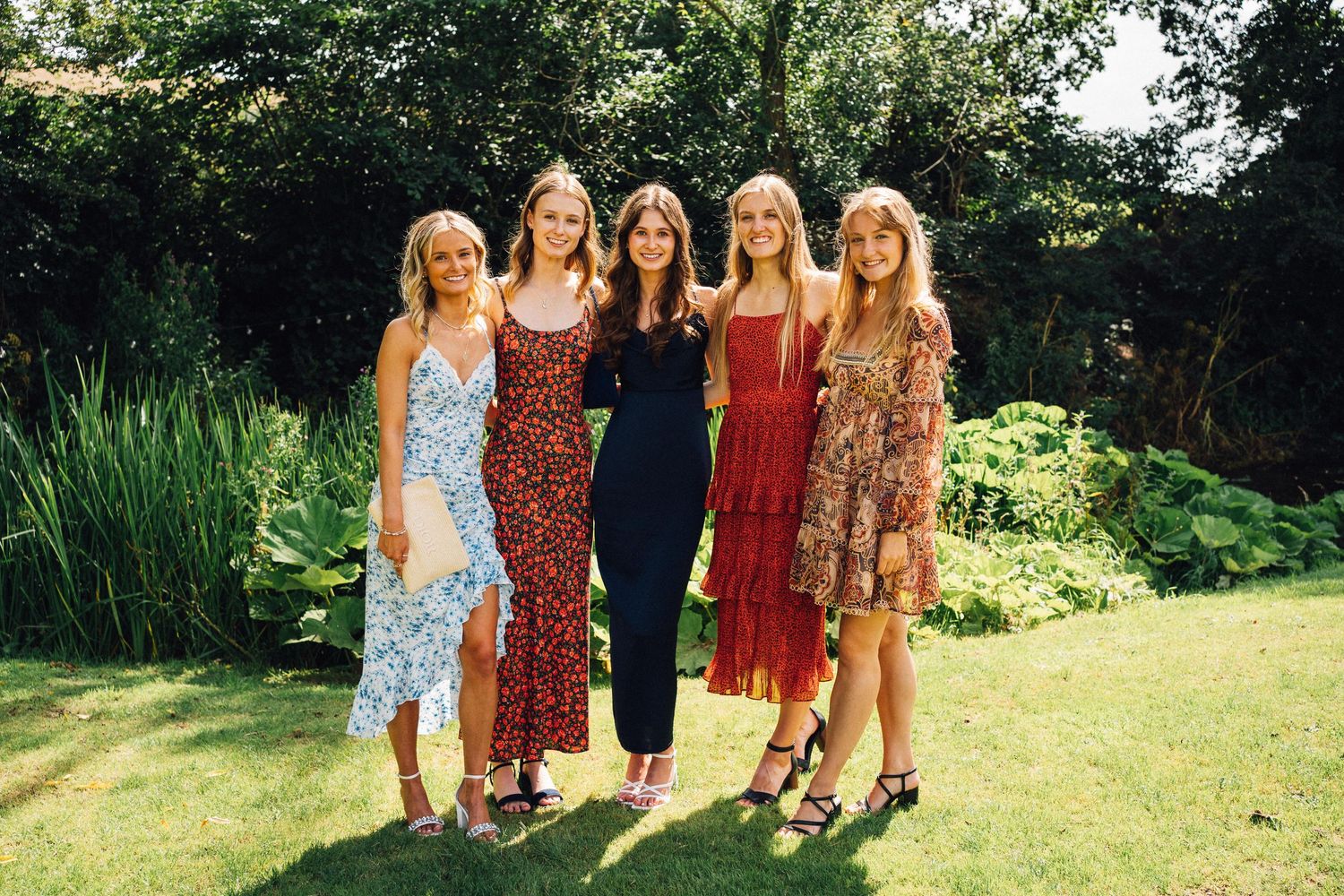 Five women in colorful summer dresses smiling together on sunny green lawn with garden backdrop.