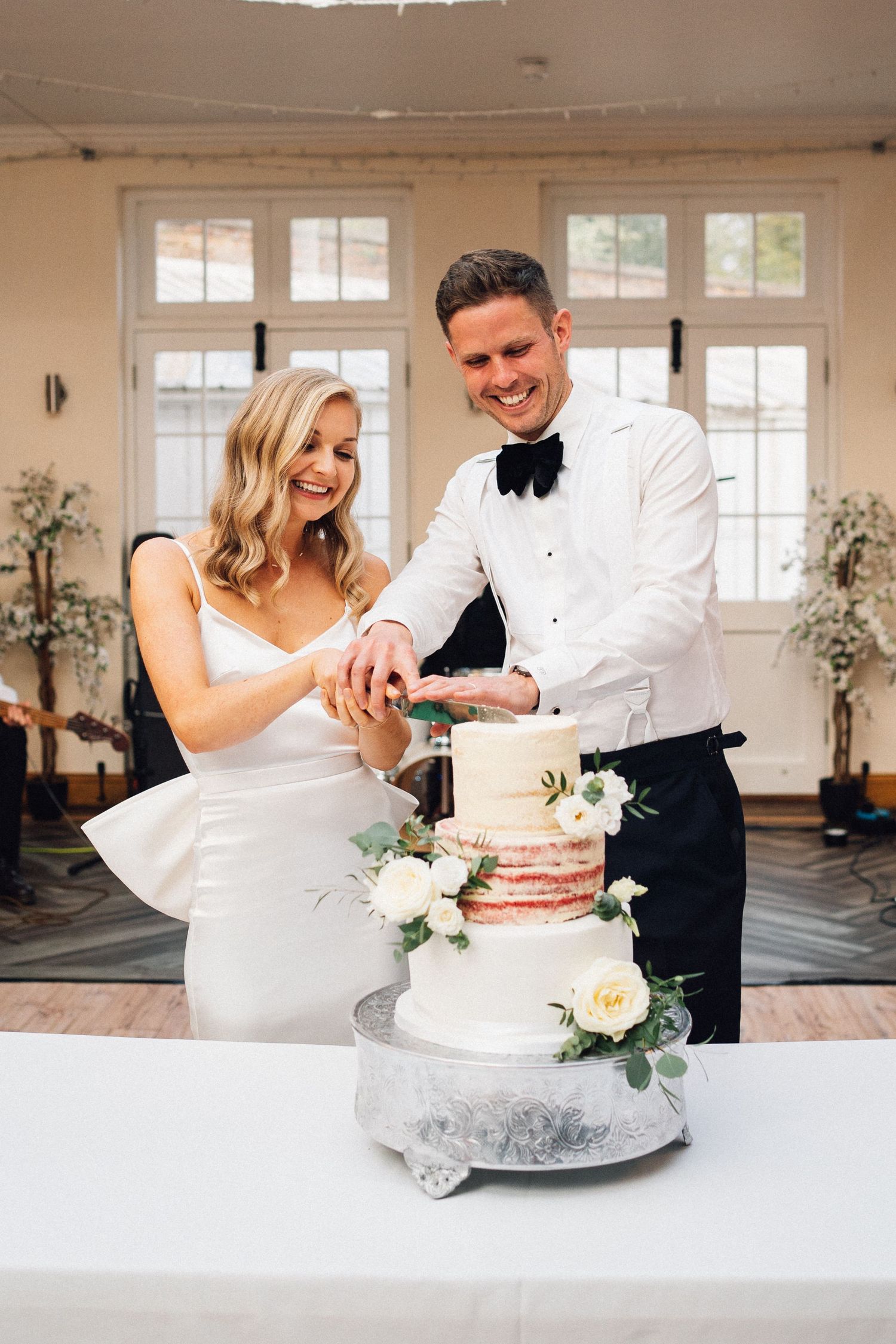 Newlyweds cutting their elegant white wedding cake together at an indoor reception.