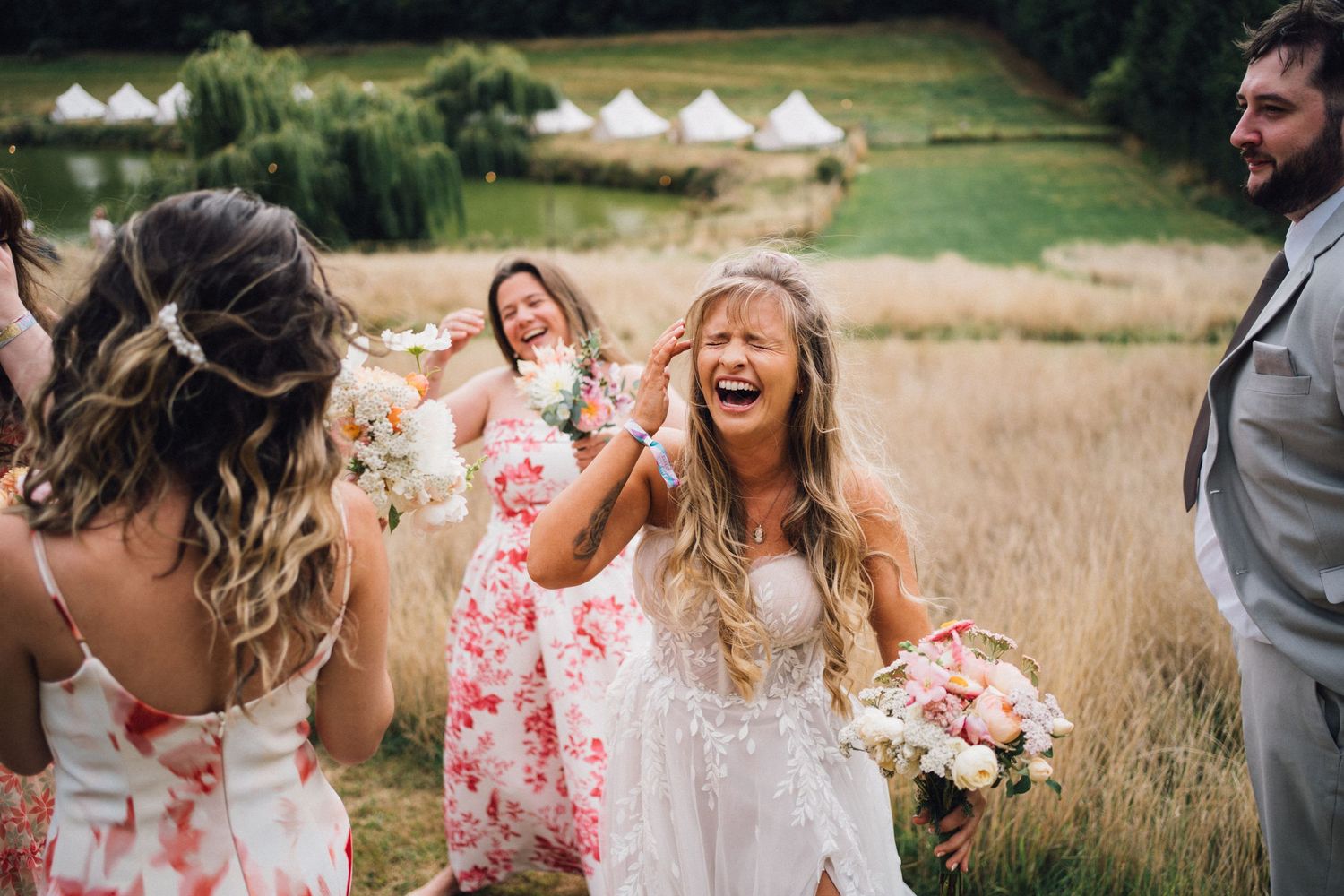 Candid moment of wedding party laughing together in a field with white tents in background.