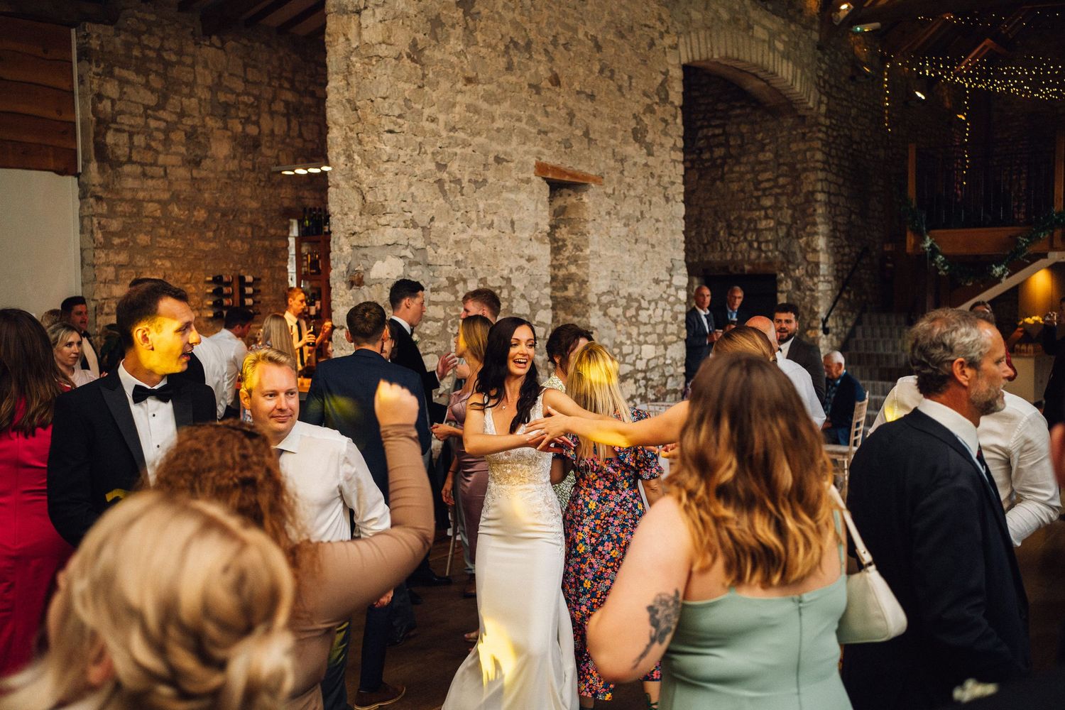Wedding guests enjoying a lively celebration in a rustic brick-walled venue with string lights overhead.
