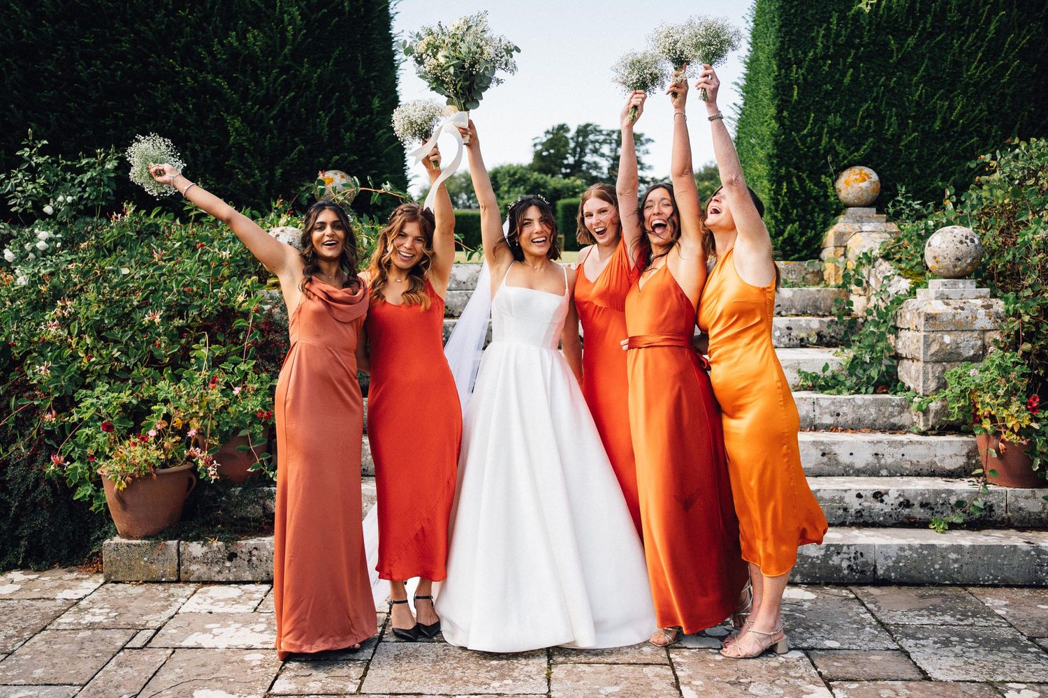 Wedding party members in orange and red dresses celebrate joyfully in a garden.