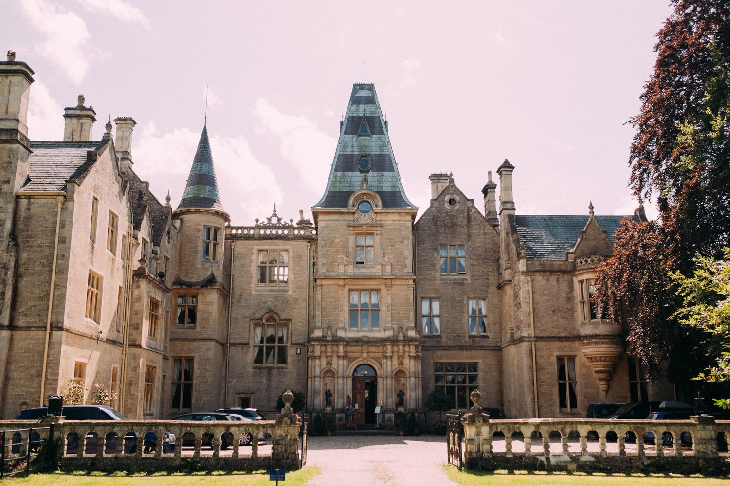 Grand Victorian Gothic manor house with ornate stone facade, pointed turrets, and manicured grounds under a cloudy sky.