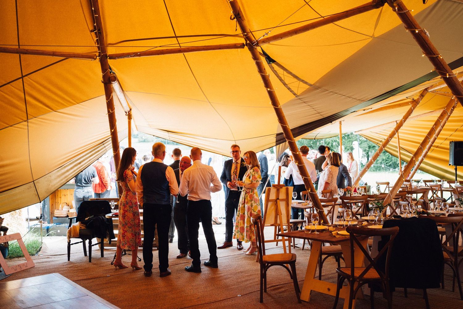 Guests mingle under a warm golden tipi tent at an outdoor wedding reception with wooden furniture.