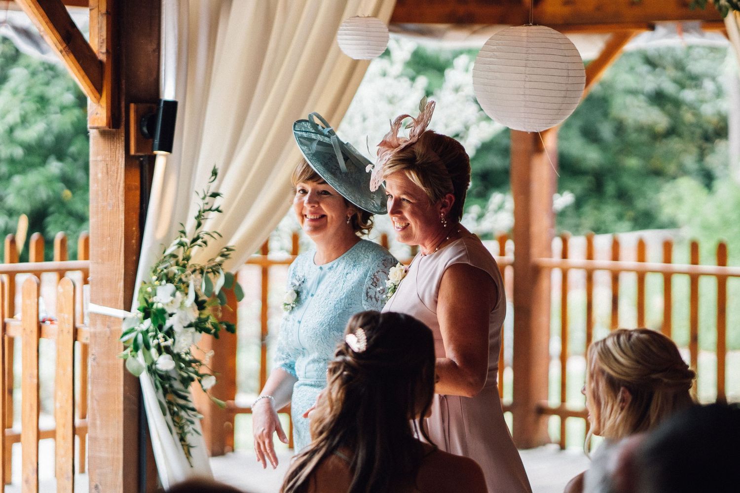 Wedding guests gather under a rustic wooden pavilion decorated with floral arrangements during an outdoor celebration.