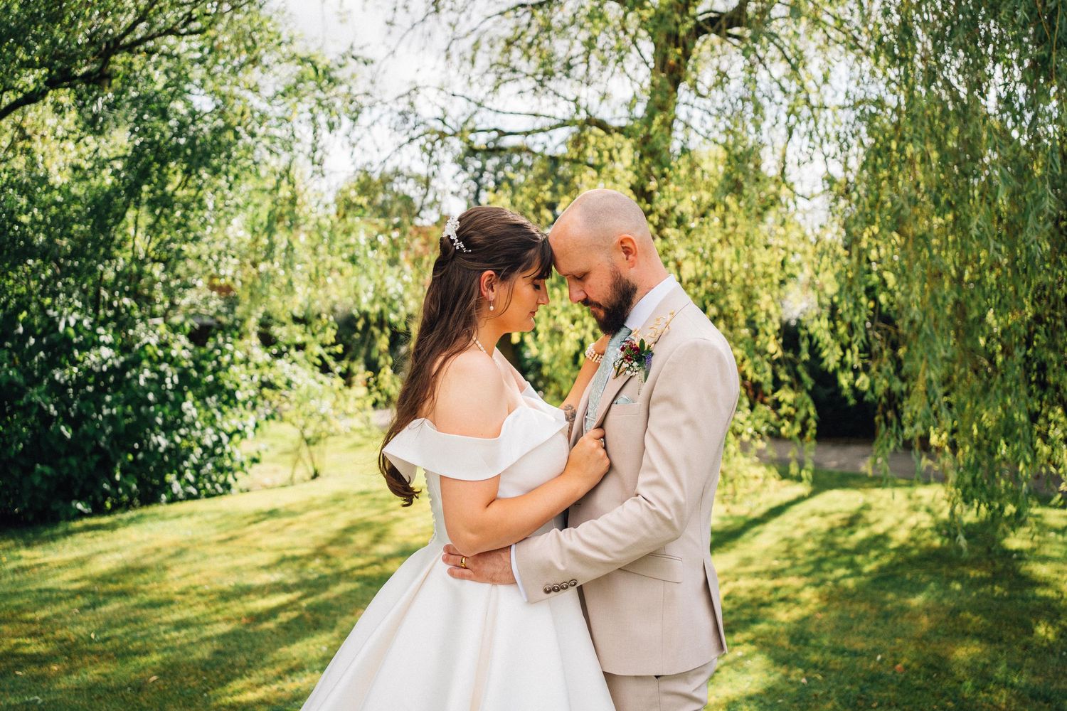 A couple shares a romantic moment under a weeping willow tree in a sunlit garden setting.