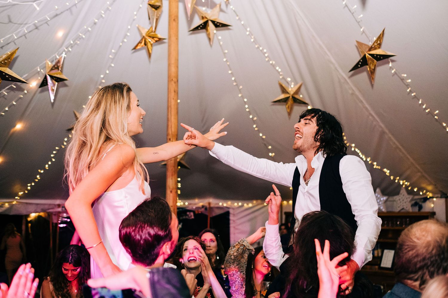 Wedding guests dance and celebrate under a tent decorated with twinkling lights and golden star ornaments.