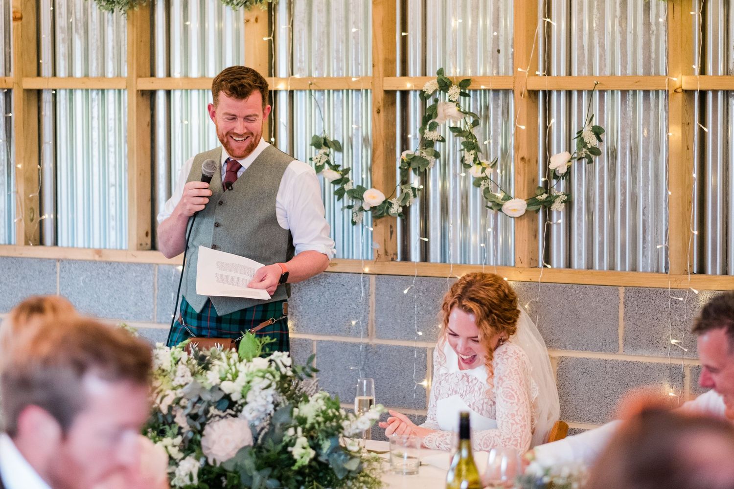 Wedding speeches taking place in a rustic barn venue with decorative greenery and lighting.