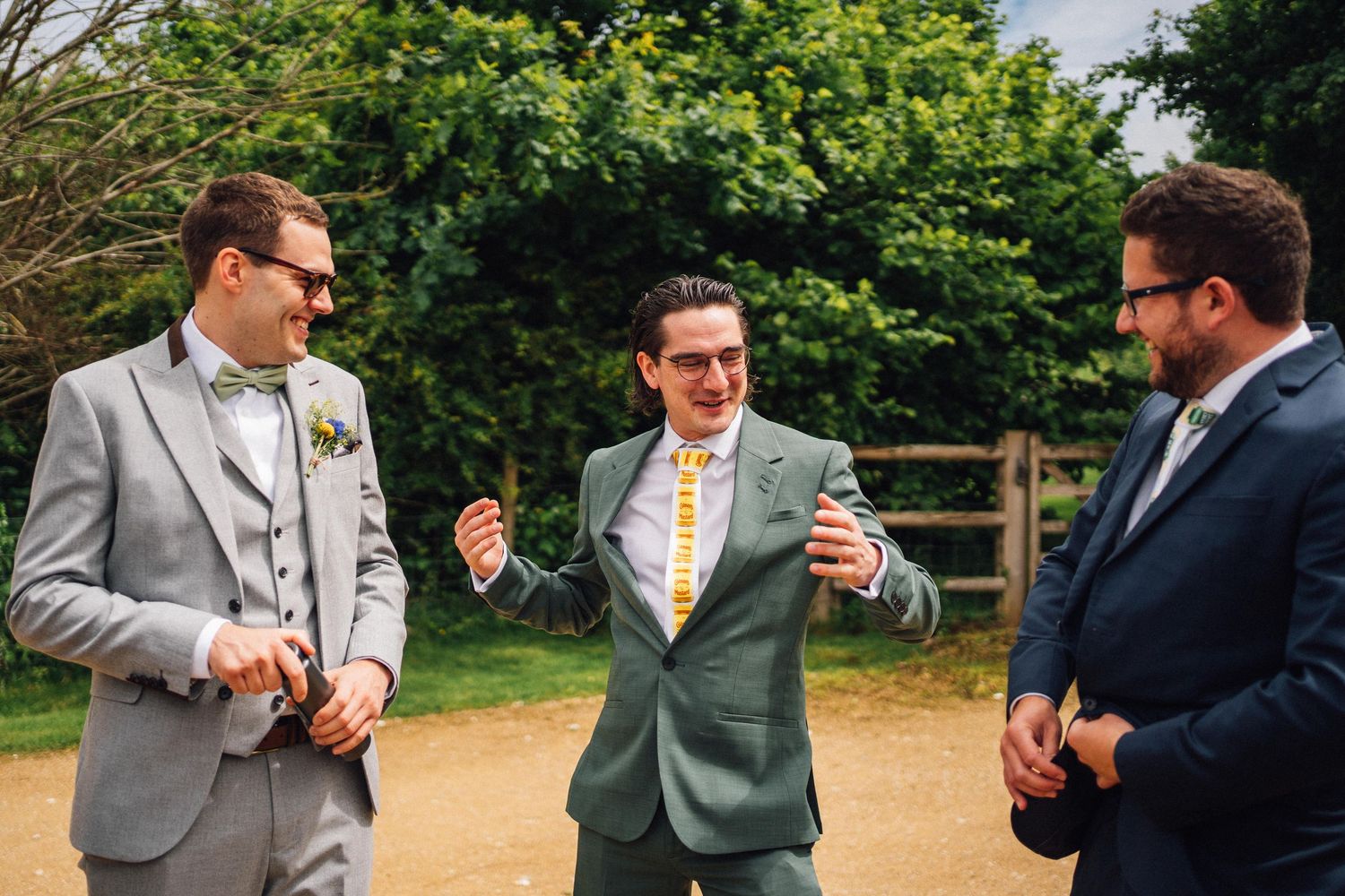 Groomsmen in formal attire engage in animated conversation while standing outdoors in a garden setting.