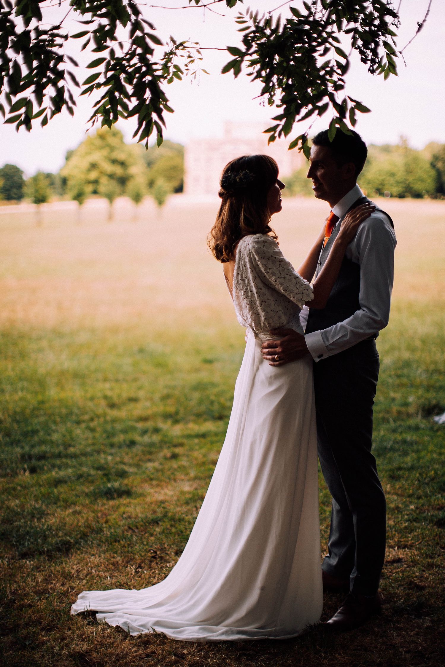 A romantic wedding couple embraces under tree branches in a sunlit field during golden hour.