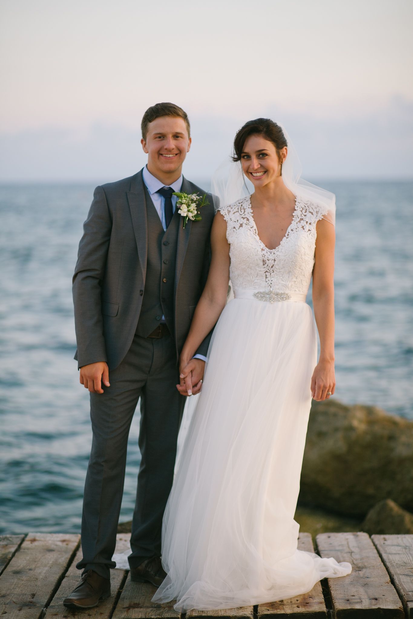 A joyful wedding portrait of newlyweds standing on a dock overlooking the ocean.