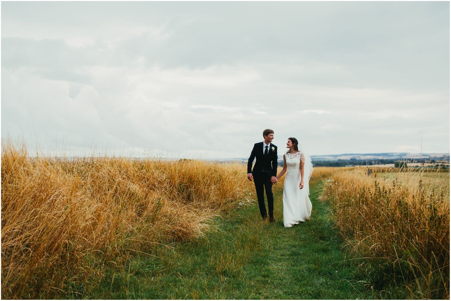 Wedding couple walking along grassy path with panoramic countryside views.