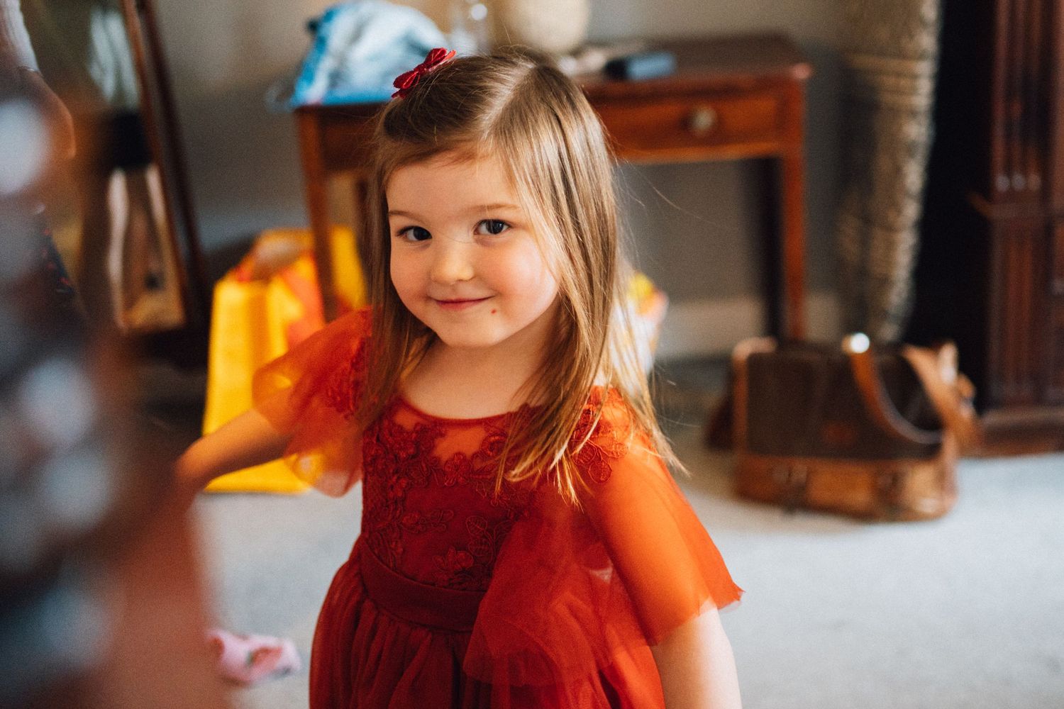A young child wearing a festive red dress smiles warmly in a cozy home setting.