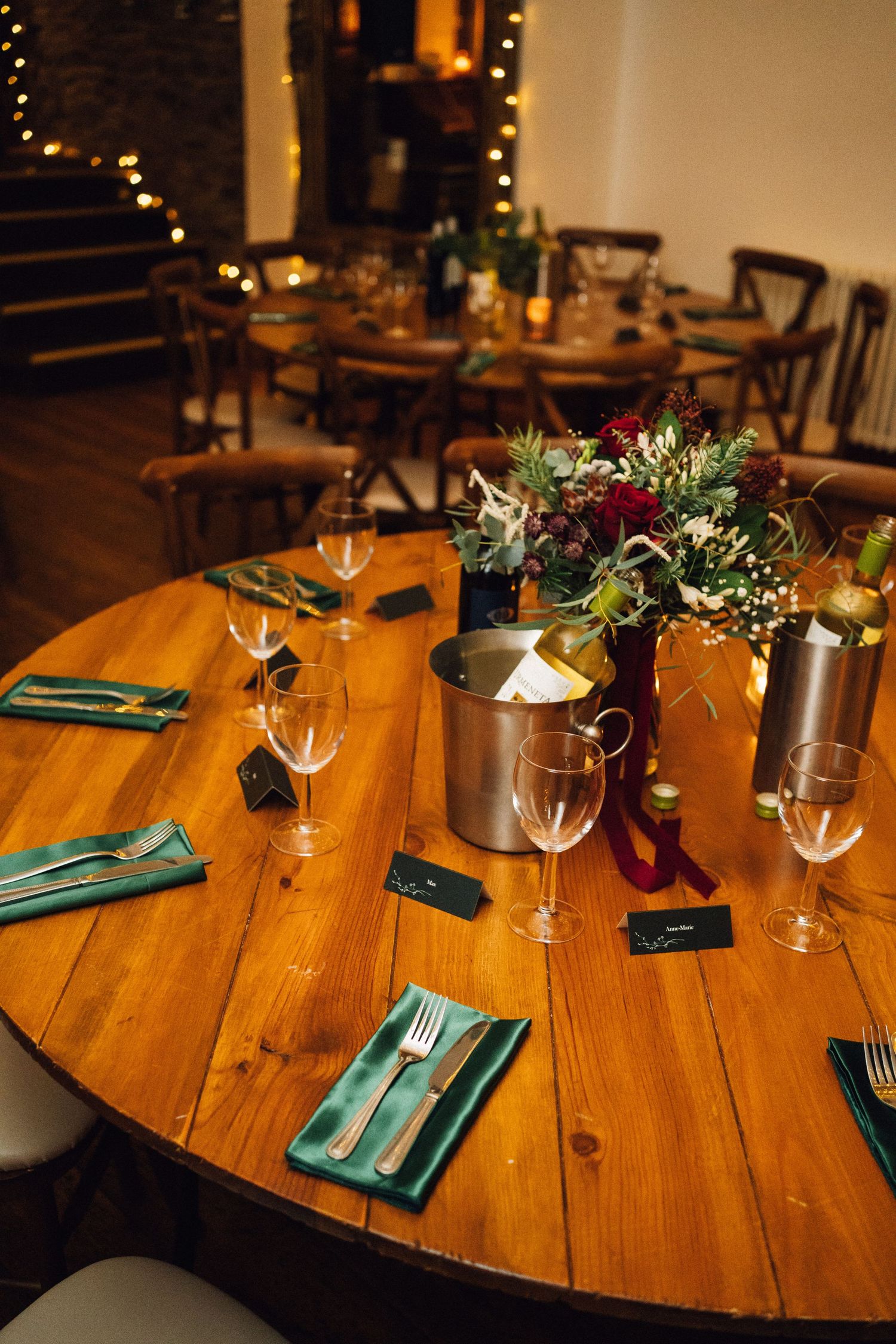 A rustic wooden round table set with turquoise napkins, wine glasses and floral centerpieces in a dimly lit restaurant.