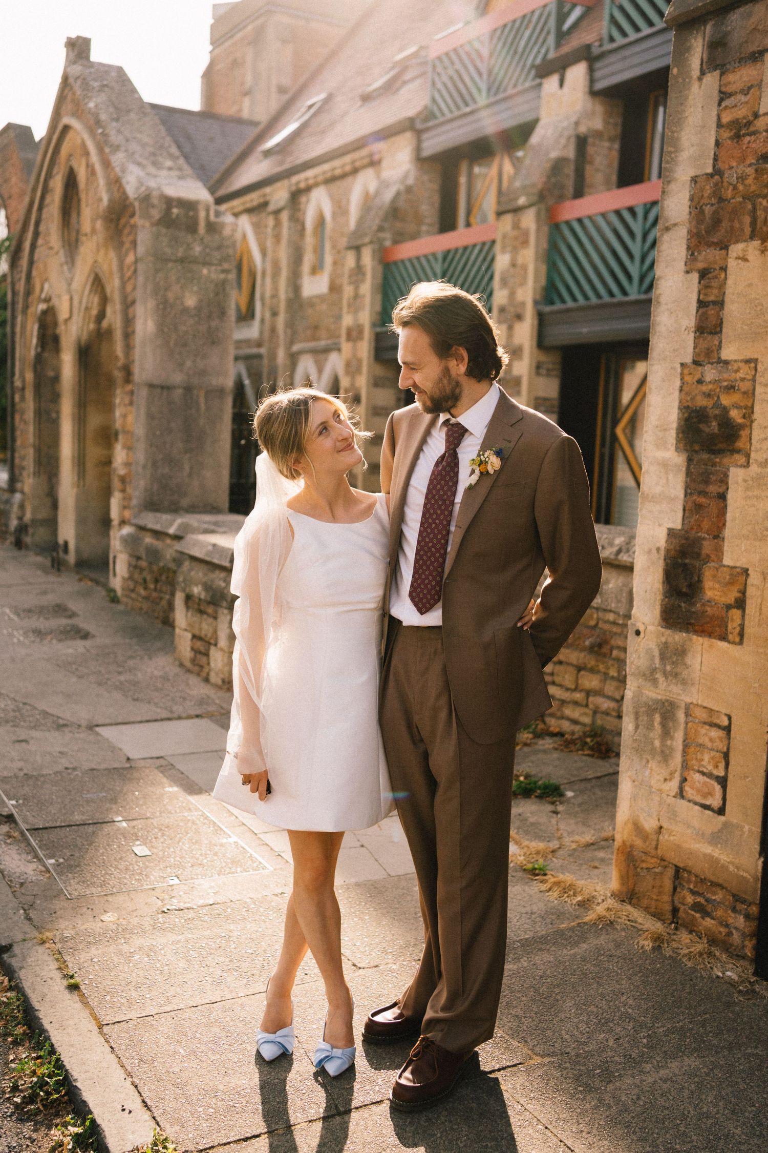 A bride in a white mini dress and groom in a brown suit share a romantic moment outside a historic stone building.