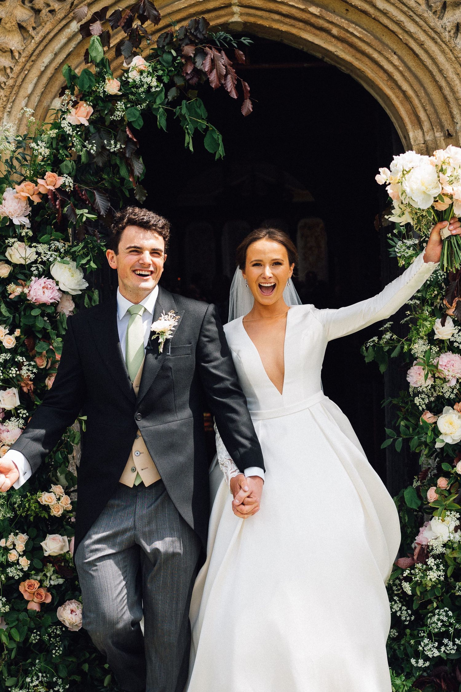 Joyful bride and groom exit a flower-adorned stone church arch, holding hands and raising arms in celebration.
