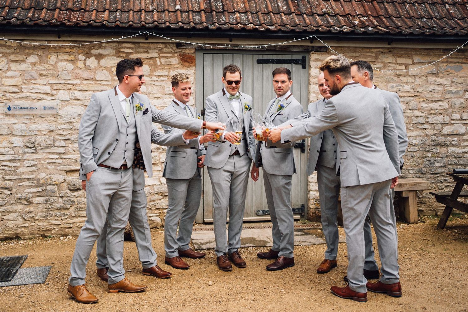 Wedding party in coordinated gray suits poses casually against stone wall backdrop.