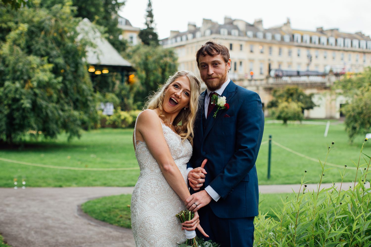 Wedding couple embraces on green lawn with historic architecture in background.