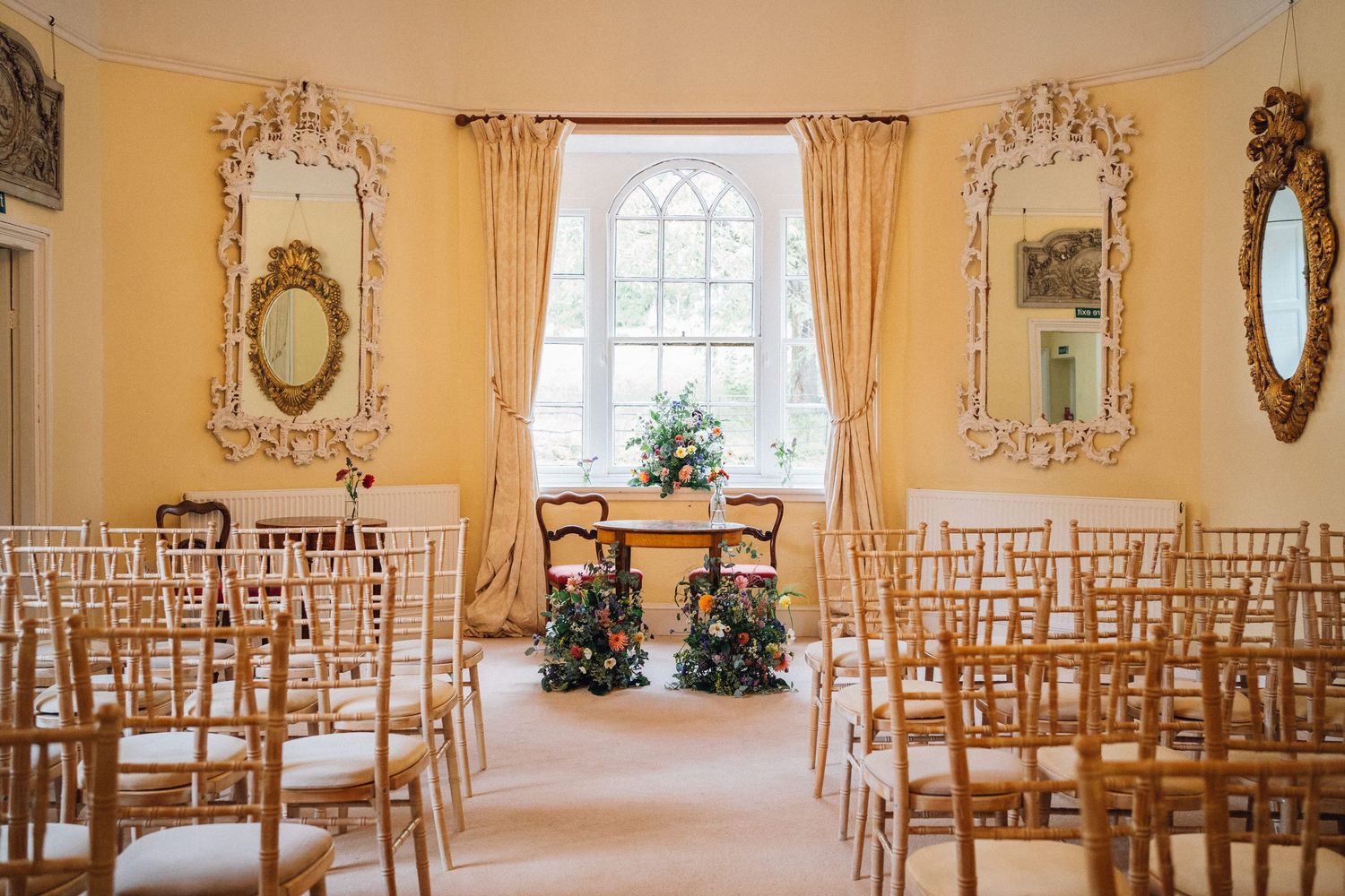 Elegant wedding venue with golden chiavari chairs and ornate mirrors in a sunlit ceremony room.