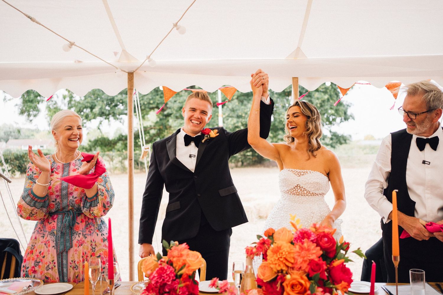 Bride and groom raise hands in celebration at wedding reception under tent with colorful floral arrangements.
