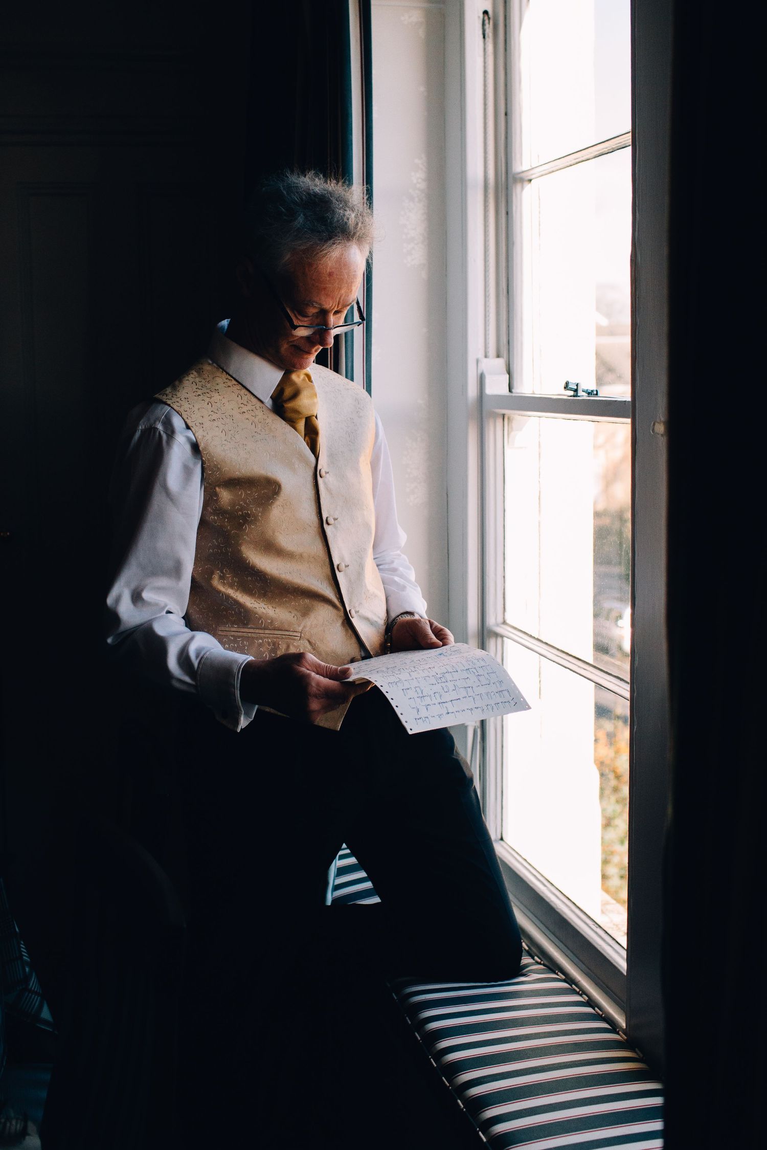 Person in formal attire reading by window light in moody atmospheric setting.