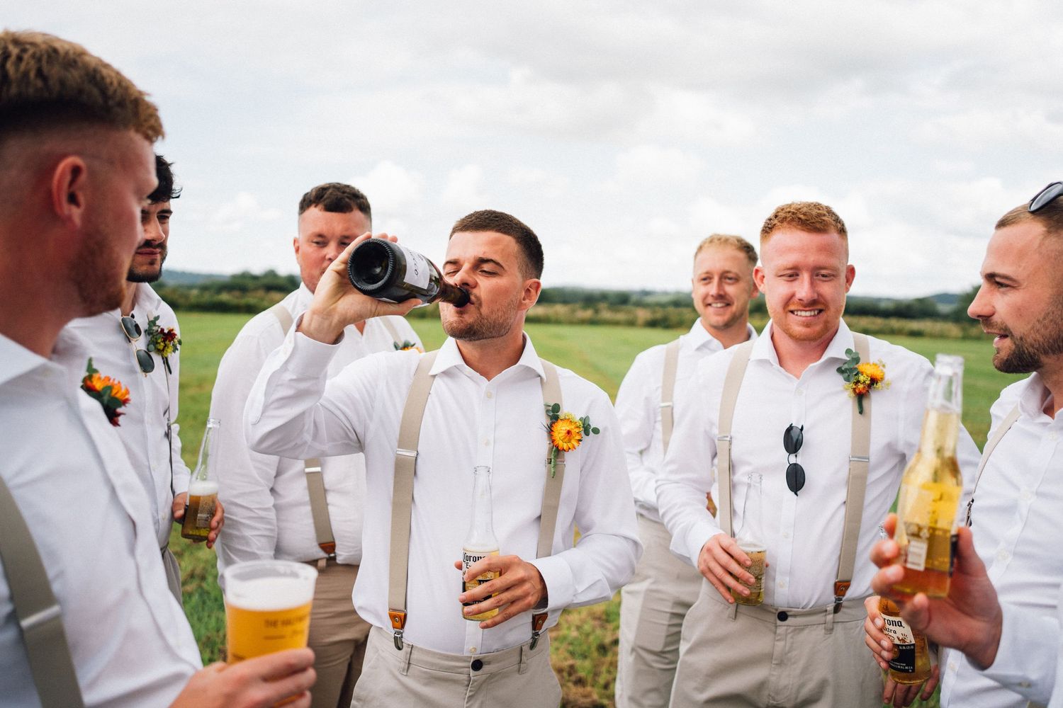 Wedding party members in white and beige attire share drinks and laughs during outdoor celebration.