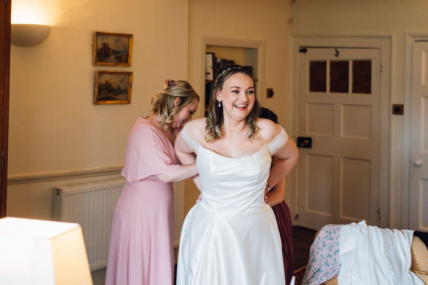 A bride in a white wedding dress gets ready with bridesmaids in pink dresses in an elegant dressing room.