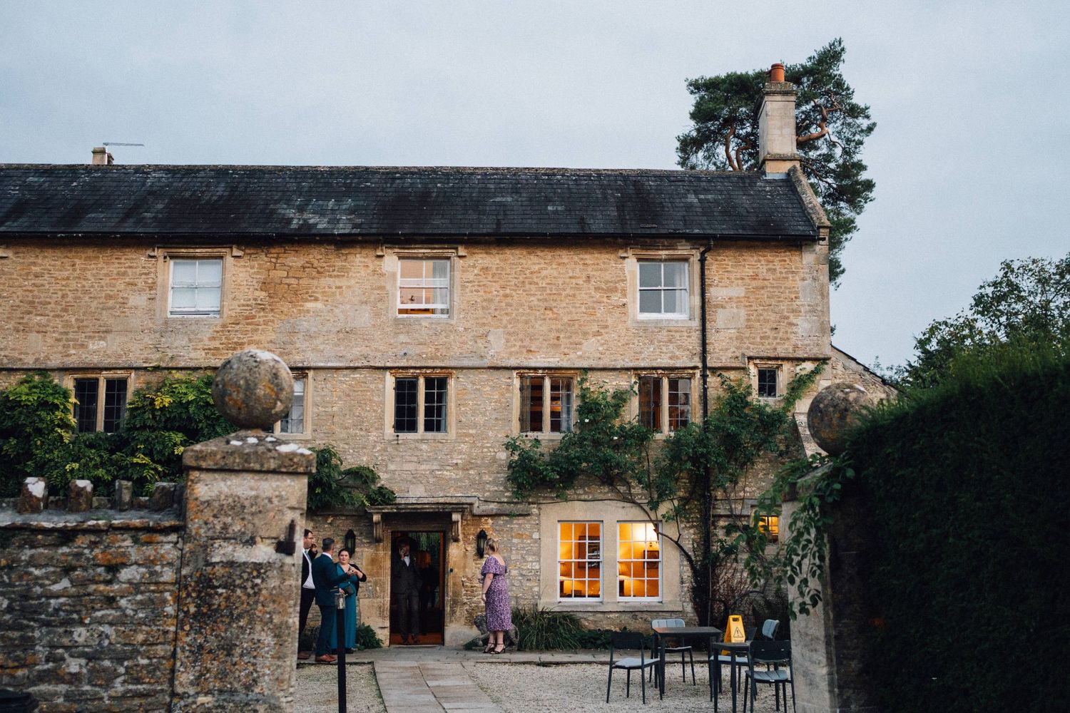 Stone cottage wedding venue with warm glowing windows and guests gathering outside at dusk.