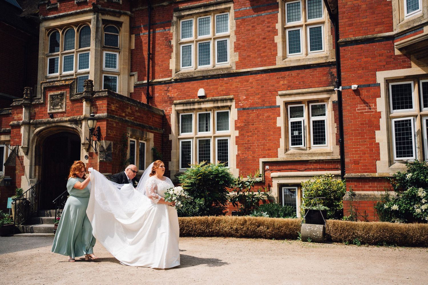 Wedding party poses in front of red brick mansion with elegant white trim and bay windows on sunny day.