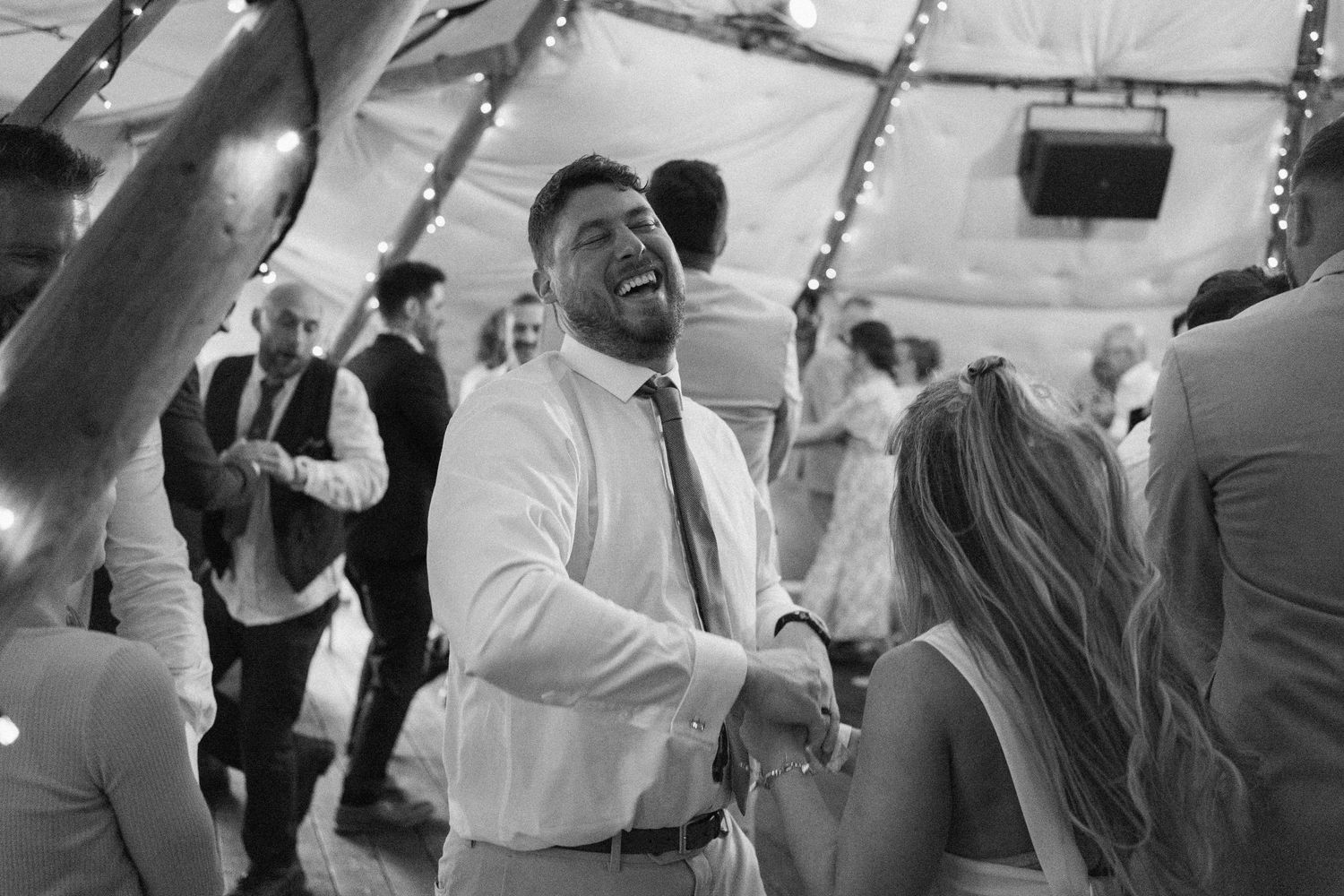 Black and white photo of guests dancing and celebrating at a wedding reception under string lights.