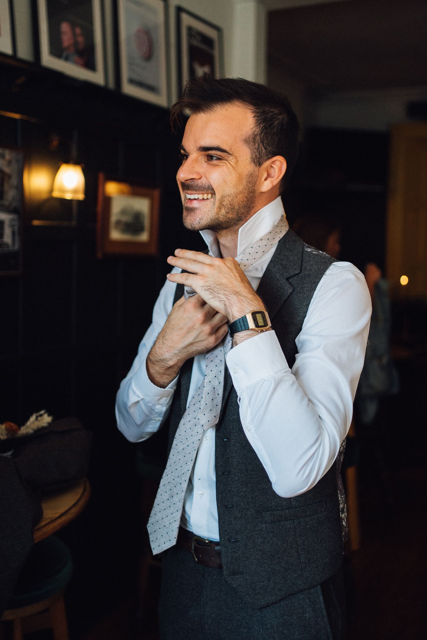 A person in a white dress shirt and grey vest adjusts their light blue patterned tie in a dimly lit room.