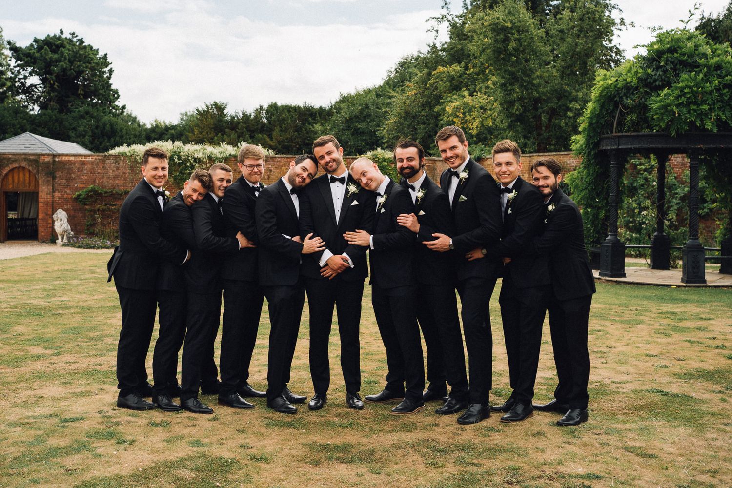 Wedding party in black suits posing playfully on grassy field with trees in background.