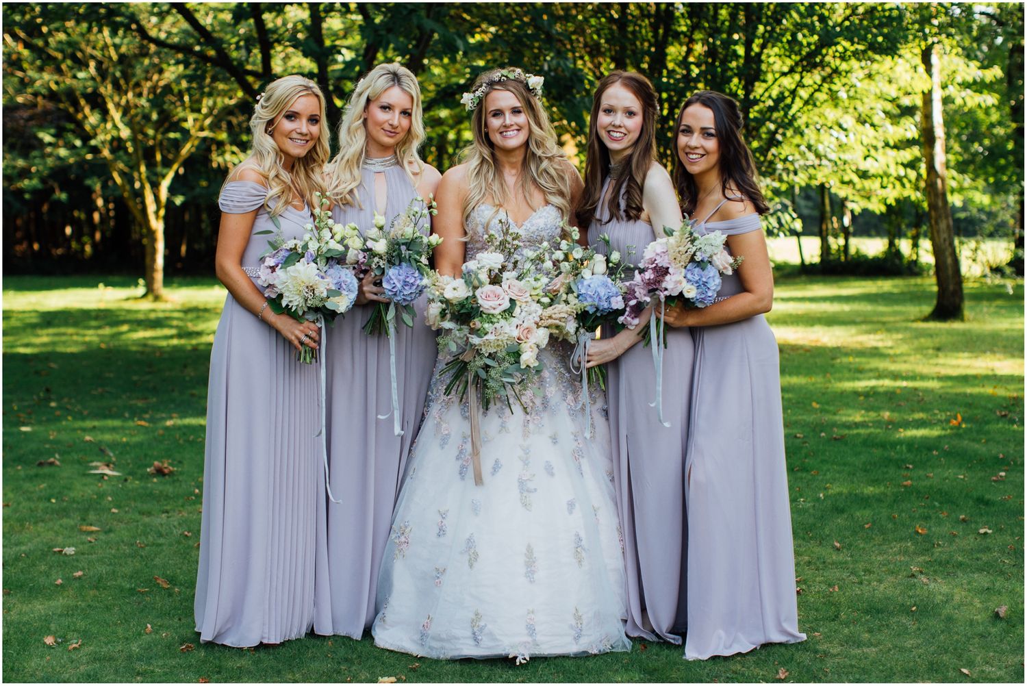 Bridesmaids in lavender dresses pose with bouquets in garden setting.