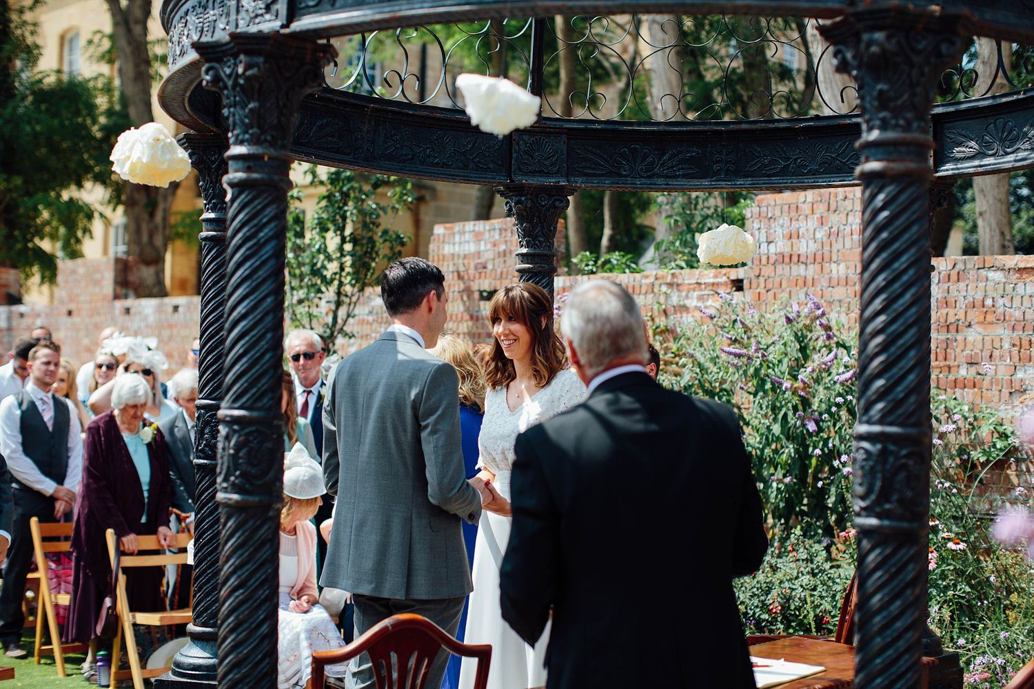 Outdoor wedding ceremony under a pergola with decorative lighting and floral arrangements.
