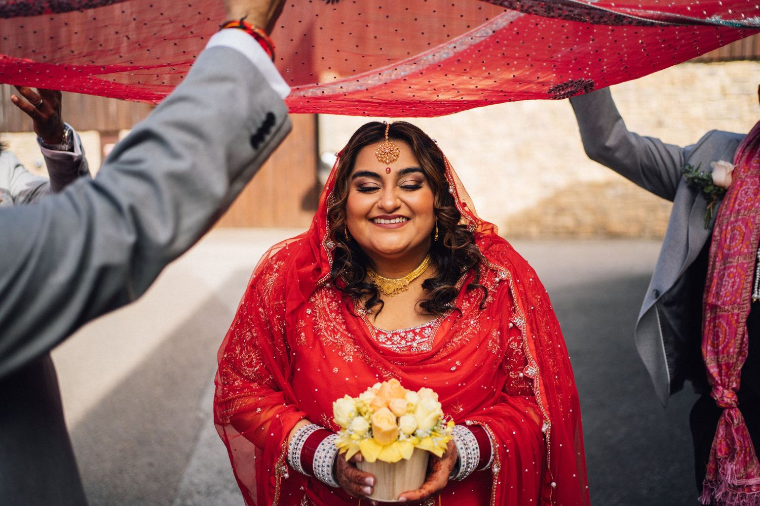 Indian bride in red lehenga smiles holding flowers under red dupatta during traditional wedding ceremony.