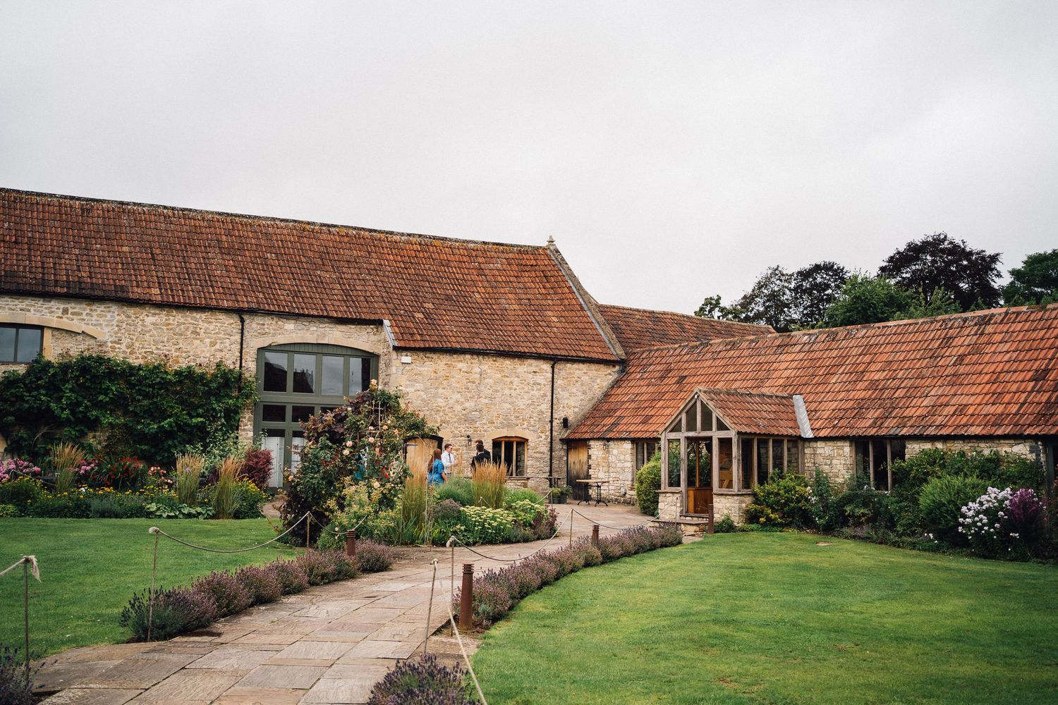 Charming stone barn wedding venue with manicured lawns, terracotta roof tiles, and colorful garden borders on an overcast day.