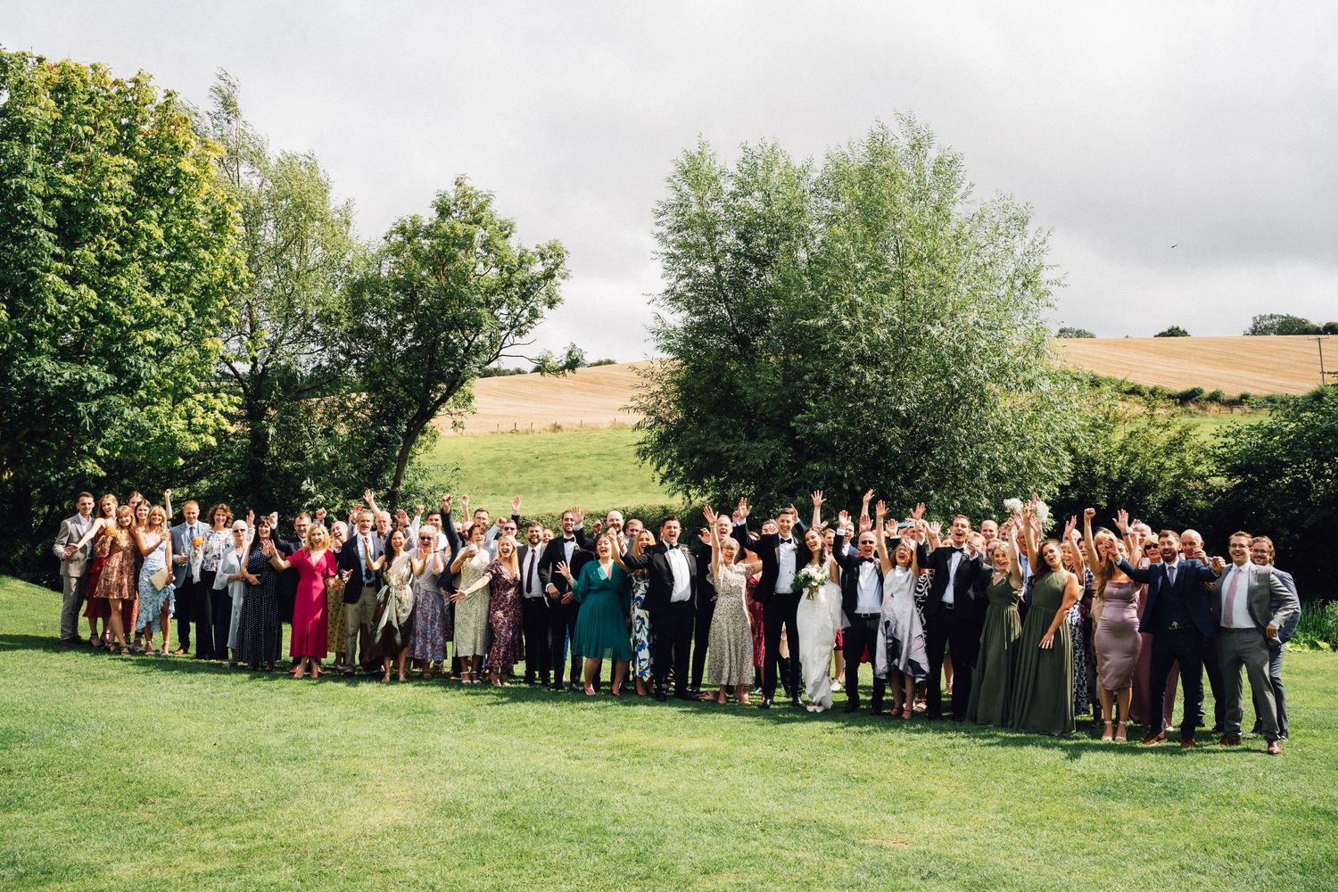 Large wedding group photo outdoors on green lawn with trees and rolling hills in background on cloudy day.