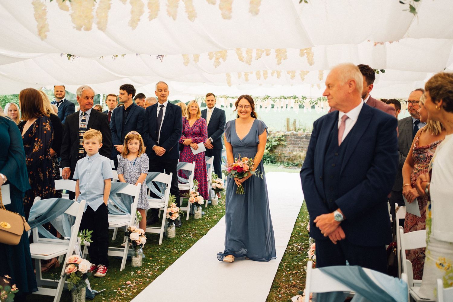 Wedding guests gather under string lights at an outdoor ceremony with attendants in blue dresses standing near white chairs.
