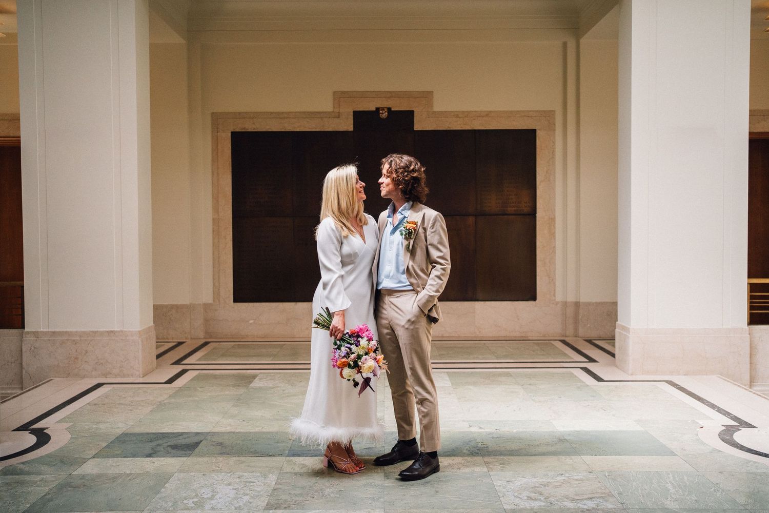 A romantic wedding couple embraces in a light-filled hallway with arched columns and stone floors.