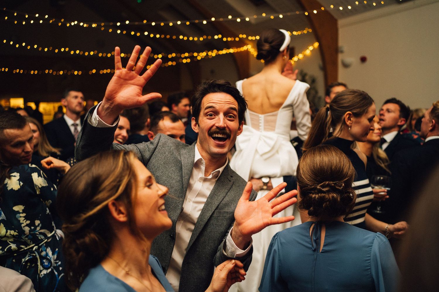 Wedding guests laugh and dance together on a dimly lit dance floor decorated with string lights above.