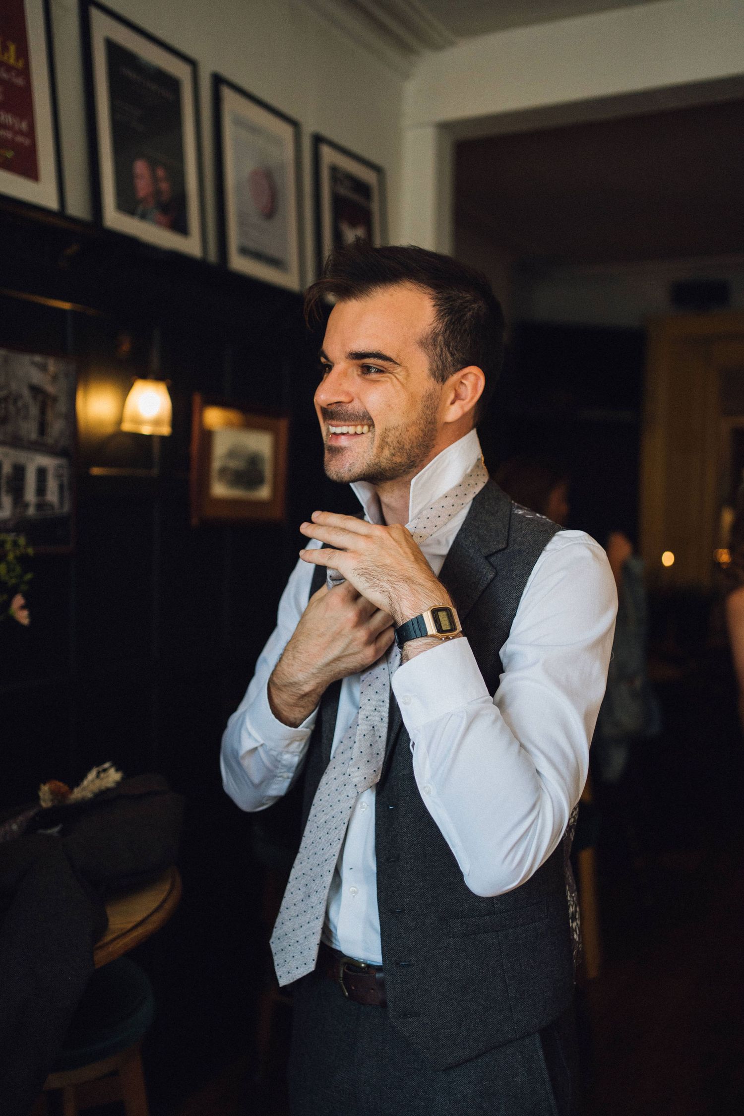 A man in a white dress shirt and gray vest adjusts his light patterned tie while smiling in a dimly lit room.