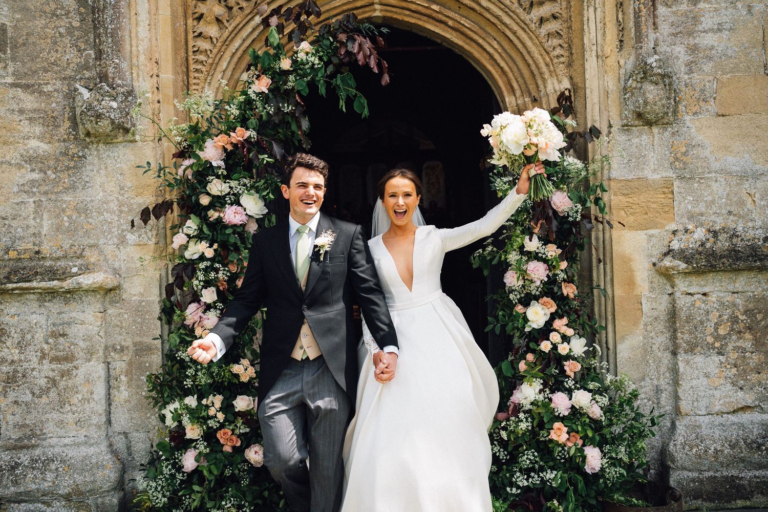 A bride and groom share joyful moments together at a historic stone church decorated with pink floral arrangements.