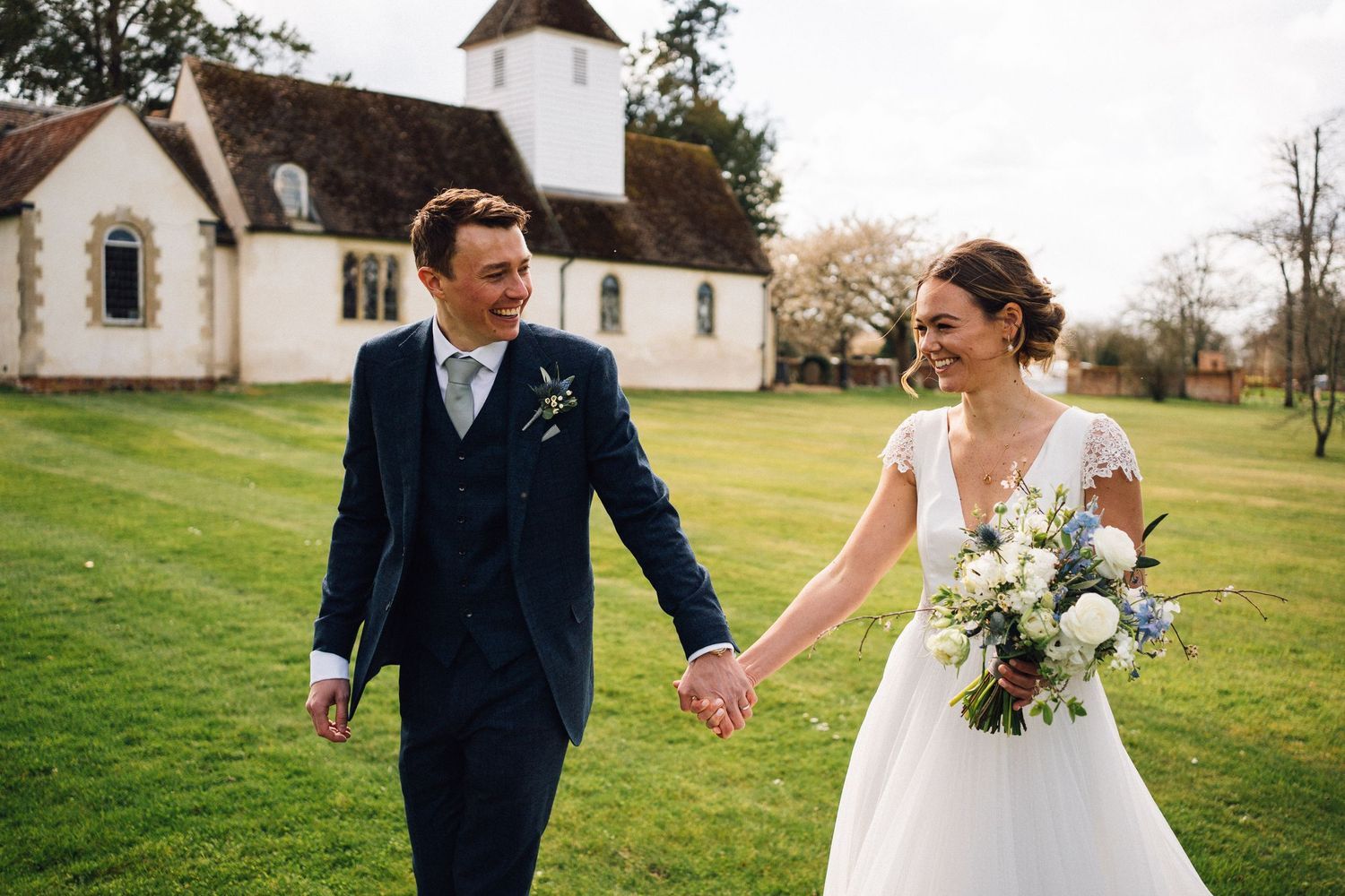 A couple in wedding attire walks hand in hand on a green lawn with a white church building in the background.