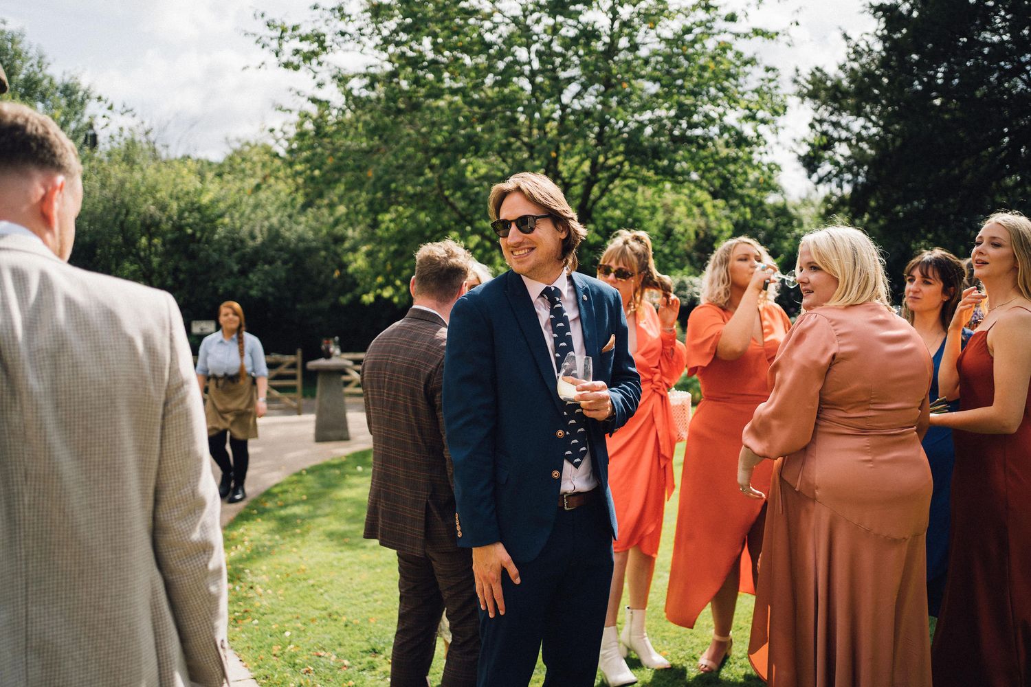 Wedding guests in formal attire gather outdoors on a sunny day, with people wearing navy suits and colorful dresses.