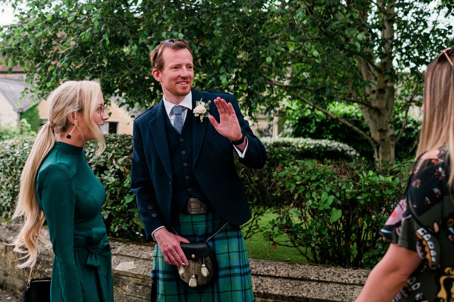 Wedding party members interacting outdoors in Scottish formal wear under trees.