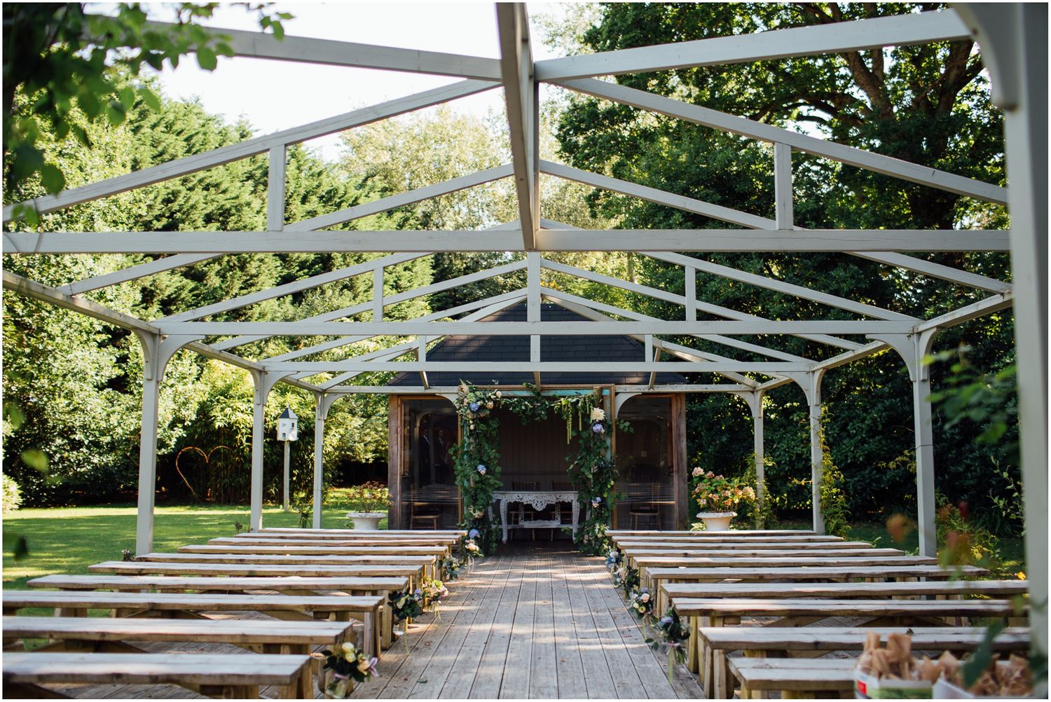 Outdoor wedding ceremony setup with white canopy and wooden benches lined up on stone path.